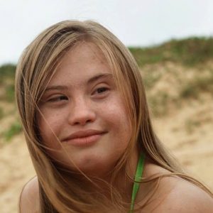 Marina, que tem síndrome de Down, deseja conhecer o mar no filme Marina não vai à praia. Na imagem, a jovem aparece na praia com um sorriso de satisfação no rosto.