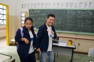 Dois adolescents com deficiência caminham de mãos dadas na sala de aula. A garota é cega e é conduzida pelo garoto que tem síndrome de Down.