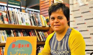 Débora Seabra, primeira professora com síndrome de Down no Brasil, posa em uma livraria ao lado do livro de fábulas escrito por ela, chamado Débora Conta Histórias.