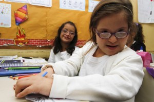 Duas garotas com síndrome de Down estão sentadas em suas carteiras em uma sala de aula inclusiva. Elas estão usando uniforma e seus materiais escolares estão sobre as carteiras. A foto ilustra o tema inclusão escolar.