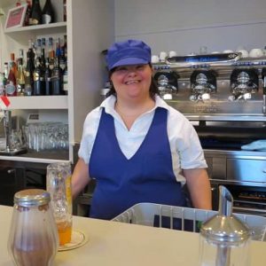 Funcionária do Milleluci Café com síndrome de Down aparece vestida com seu uniforme de trabalho e em frente à máquina de café. Ela sorri e aparece feliz em estar ali.