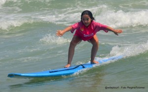 Letícia tem síndrome de Down. Na foto, ela aparece surfando em uma prancha azul no mar. Em pé, Letícia se equilibra para surfar na onda. Com o surf, ela aprendeu que pode superar qualquer desafio.