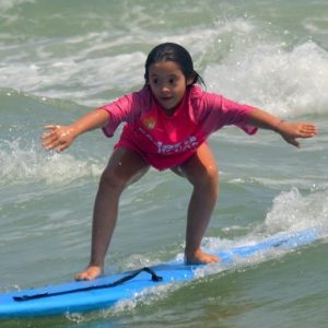 Letícia tem síndrome de Down. Na foto, ela aparece surfando em uma prancha azul no mar. Em pé, Letícia se equilibra para surfar na onda.