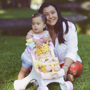 Gisele e sua filha Luisa, que tem síndrome de Down, aparecem na foto. Luisa é uma bebê ainda, está em pé e segura uma cadeirinha de brinquedo. Gisele está sentada atrás da filha. As duas estão na grama.