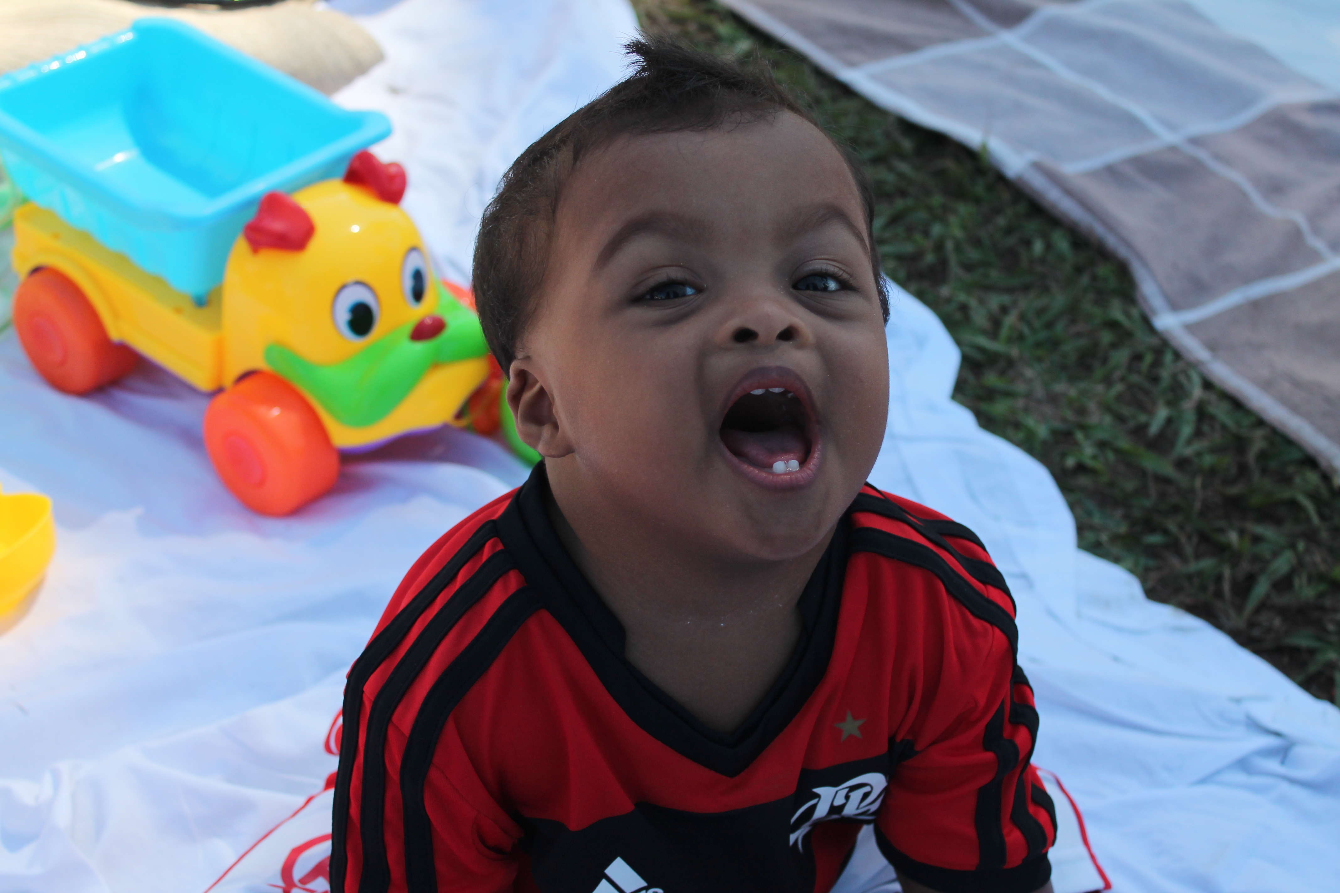 Bebê negro com síndrome de Down. Ele está em cima de uma toalha de piquenique com alguns brinquedos em volta e abre a boca tentando falar. Essa imagem ilustra o exercício de estimular a fala.