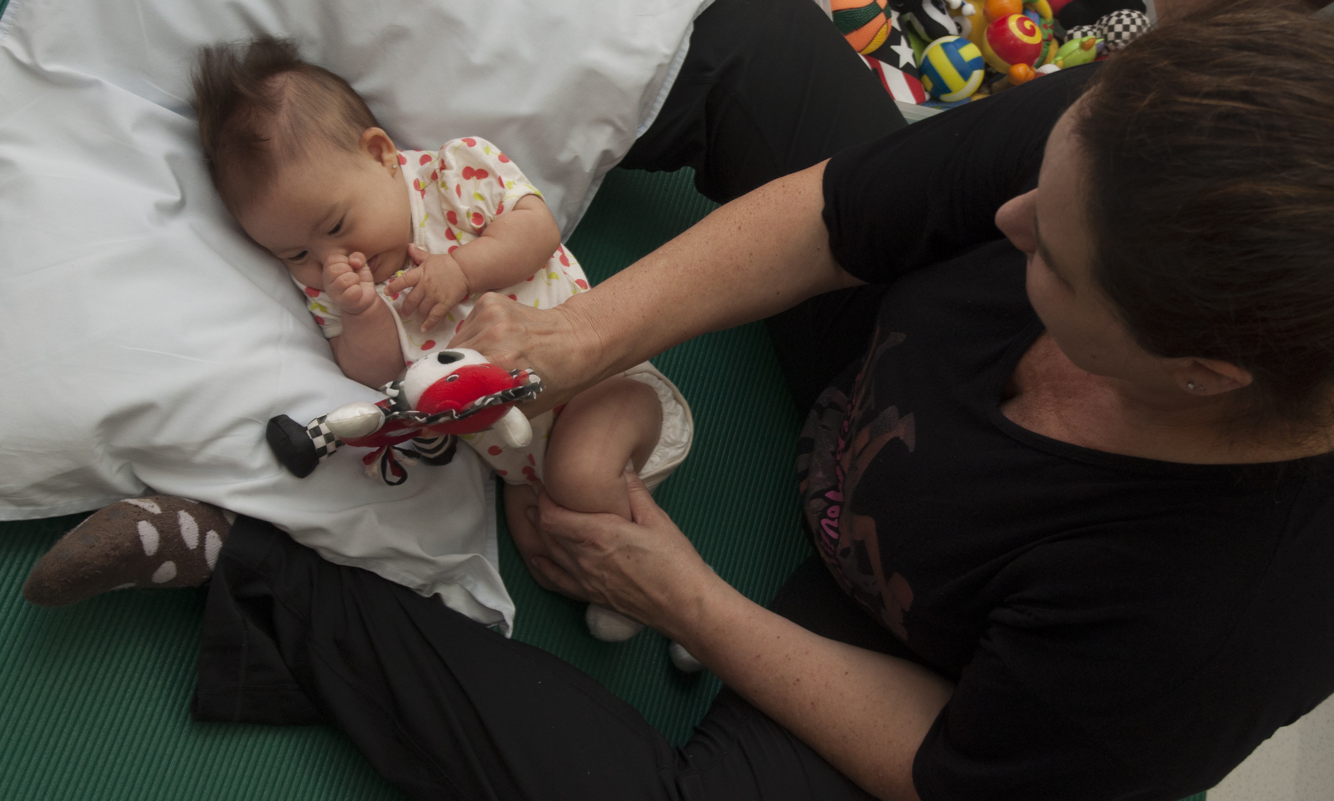 Um bebê com síndrome de Down faz exercícios para estimular a sua coordenação motora. Ele está deitado em um travesseiro e segura um bichinho de pelúcia. Uma mulher sentada no chão e na frente do bebê segura o sei corpo de lado para ajudá-lo nos exercícios de estimulação.