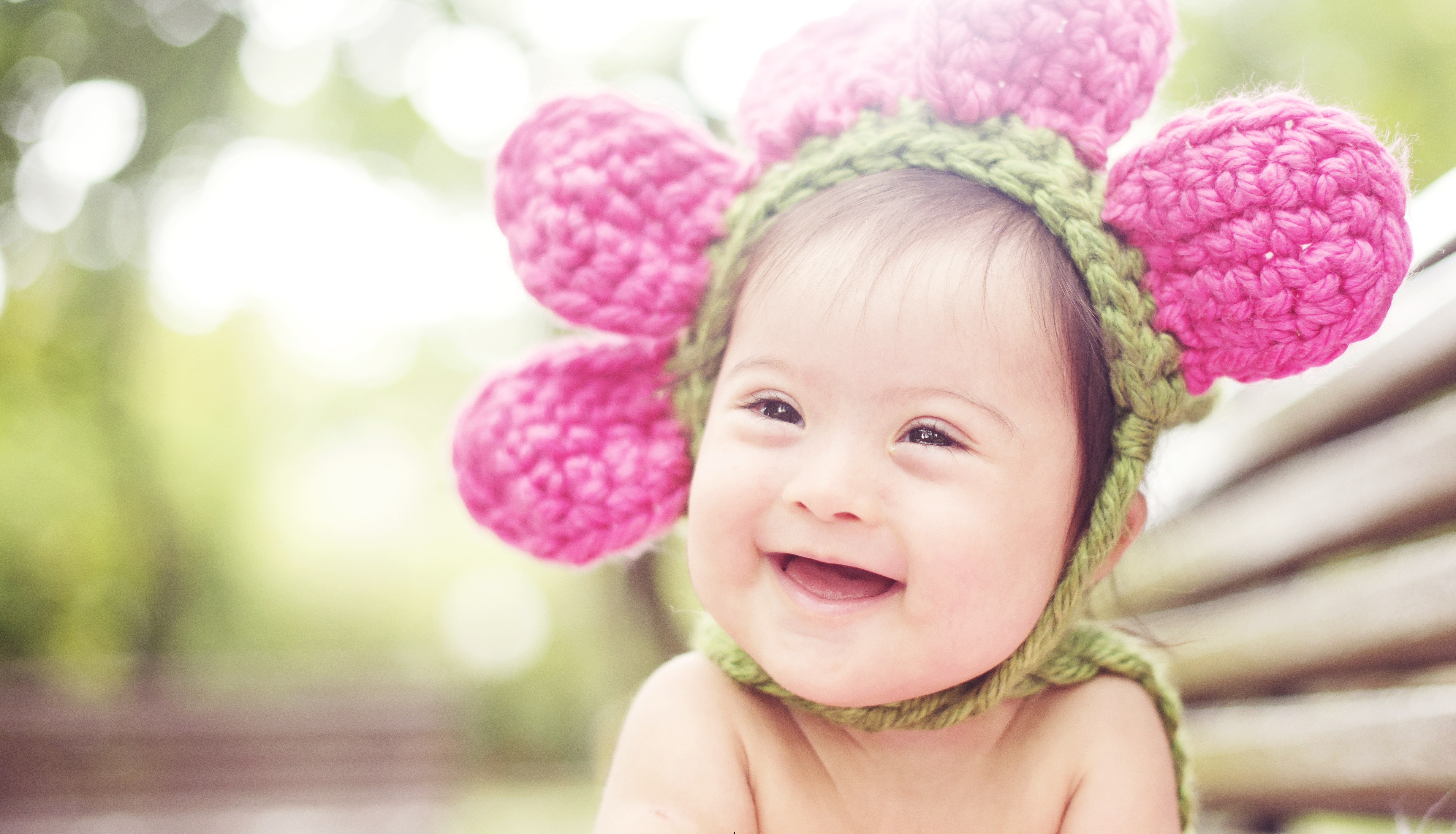Um bebê com síndrome de Down ri para a foto. Ele está muito feliz. Ele usa um chapéu em forma de flor na cabeça.