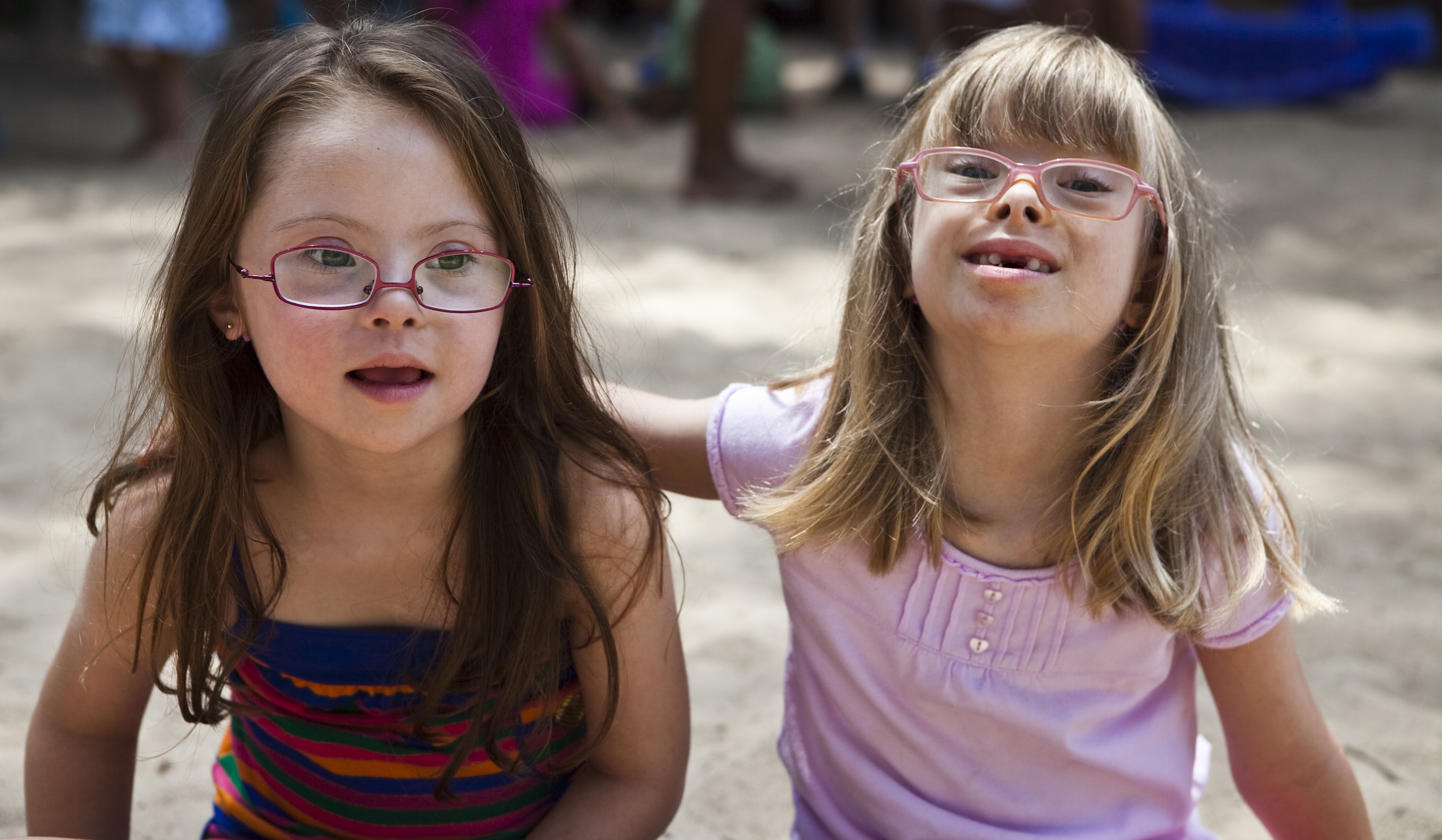 Duas garotas com síndrome de Down estão sentadas de frente para a foto. A que está do lado esquerdo olha levemente para o lado, enquanto a que está do lado direito olha para frente e apoia o braço nas costas da amiga.