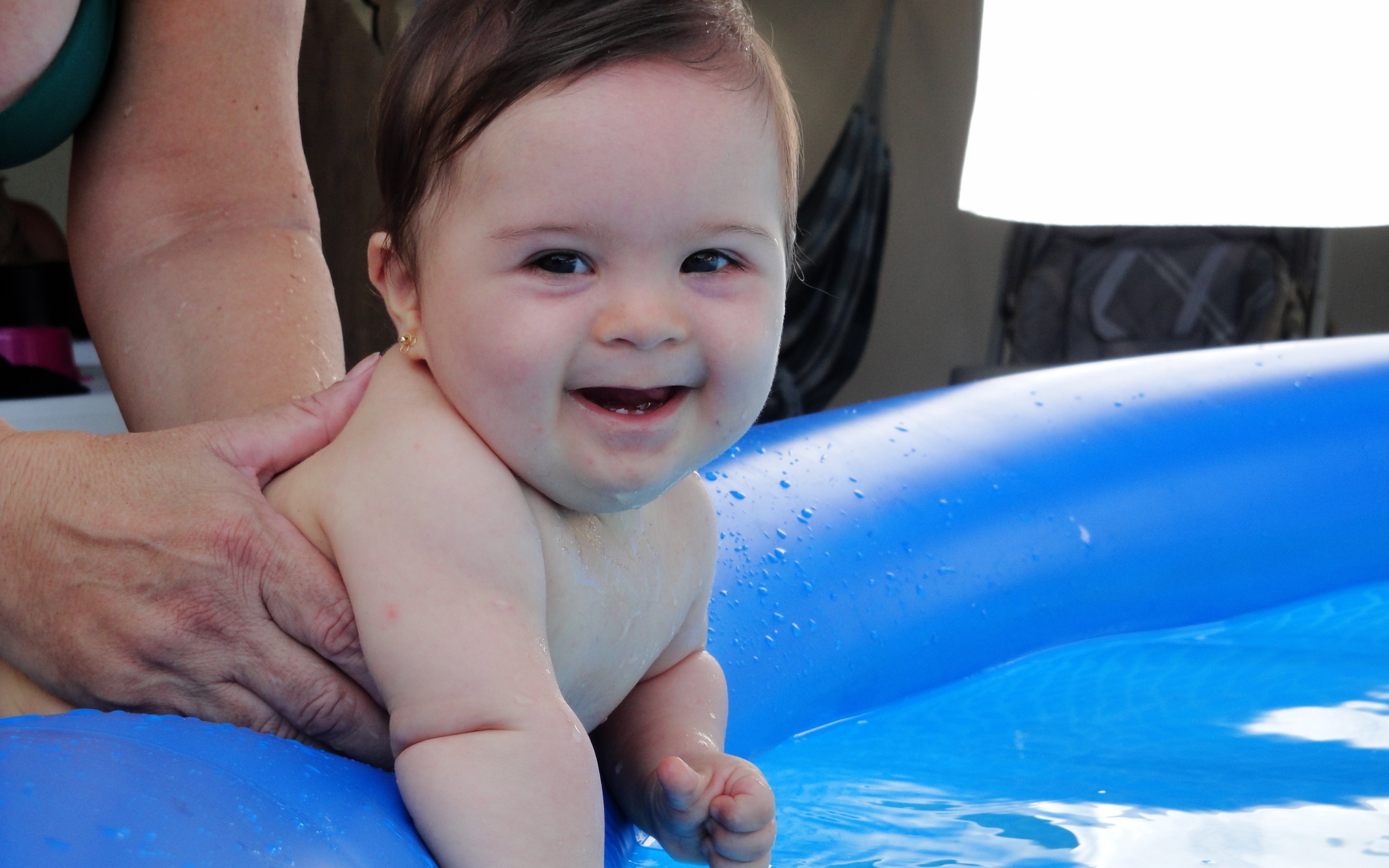 Bebê branca com síndrome de Down fazendo estimulação em uma piscina inflável. As mãos de alguém a seguram pelo corpo e a menininha sorri para a câmera.