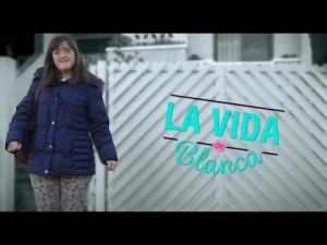 Imagem do vídeo La Vida de Branca. Na imagem, Blanca, uma jovem com síndrome de Down, aparece em pé em frente ao portão de sua casa, como se estivesse mostrando aonde mora. O nome A Vida de Blanca aparece sobre a imagem, no espaço do portão.